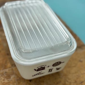 Puede incluir: Recipiente rectangular Pyrex blanquecino con tapa de vidrio transparente y acanalado. El plato presenta ilustraciones marrones de artículos de cocina. La tapa transparente permite ver el contenido. Para el almacenamiento de alimentos.