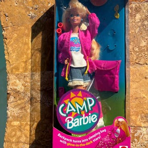 May include: A vintage Camp Barbie doll in its original packaging. The doll wears a pink jacket, white shirt, and denim shorts. The packaging features the text "Camp Barbie" and "Backpack turns into sleeping bag with glow-in-the-dark stars!"