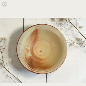 May include: A round ceramic bowl with a cream-colored interior featuring a swirling, reddish-brown glaze pattern. The exterior rim shows a dark brown tone, and the bowl has a smooth, circular shape with a visible depth and a wide, open top.