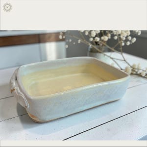 Puede incluir: Una fuente de horno rectangular de cerámica con un esmalte brillante de color crema y sutiles tonos amarillos. El recipiente cuenta con dos asas integradas en los laterales con un acabado decorativo texturizado y una base interior lisa.