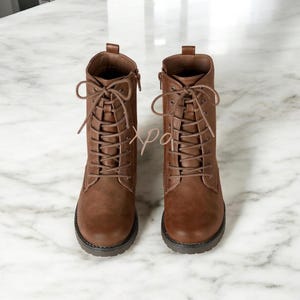 May include: A pair of brown leather combat boots. The boots feature a lace-up design with multiple eyelets and a side zip. The soles are dark brown, and the boots are set against a marble background.