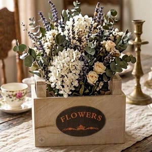 Puede incluir: Una caja de madera llena de una variedad de flores, incluyendo hortensias blancas, lavanda y rosas color crema. La caja tiene una etiqueta ovalada que dice "FLOWERS" en letras naranjas. También se ven una taza de té y un candelabro.