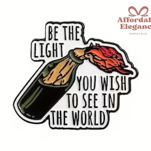 Könnte beinhalten: Schwarz-weißer Emaille-Pin mit einer Flasche und einem brennenden Docht. Der Pin zeigt den Text "BE THE LIGHT YOU WISH TO SEE IN THE WORLD". Der Pin hat einen schwarzen Rand und einen weißen Hintergrund.