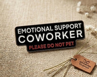 May include: A rectangular black enamel pin with white text that reads EMOTIONAL SUPPORT COWORKER and red text below that reads PLEASE DO NOT PET. The pin has a metallic border and is displayed alongside a small brown leather tag.