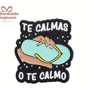 Op de afbeelding: Zwarte pin met de tekst "TE CALMAS O TE CALMO" in het wit. De pin toont een hand die een lichtblauwe slipper met een gele band vasthoudt. De pin heeft een zwarte rand met witte stippen.
