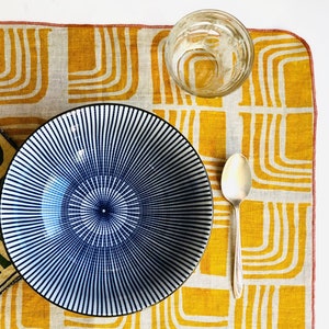 Op de afbeelding: Een placemat van linnen met een geel en groen patroon, een blauwe en witte gestreepte kom, een glas en een zilveren lepel.