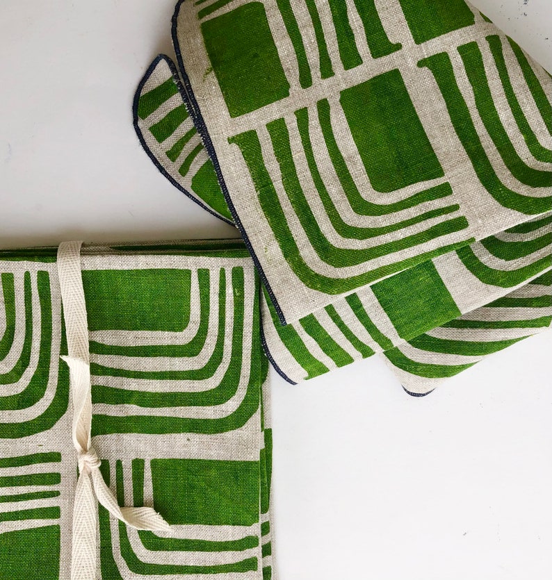 Pode incluir: Conjunto de quatro guardanapos de linho verde com um padr&atilde;o geom&eacute;trico branco. Os guardanapos est&atilde;o dobrados e amarrados com uma fita branca.