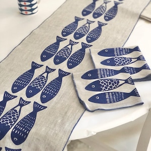 May include: A linen table runner with a blue and white fish design. The runner is on a white table with a blue and white mug.