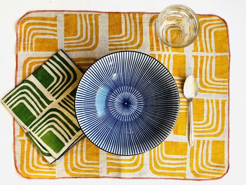 Op de afbeelding: Een placemat van linnen met een geel en groen patroon, een blauwe en witte gestreepte kom, een glas en een zilveren lepel.