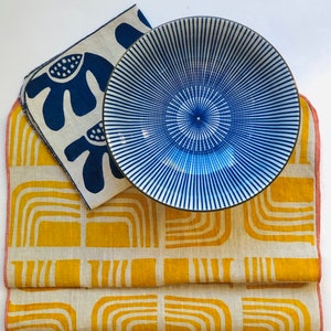 Op de afbeelding: Een set van twee linnen servetten met geometrische patronen. Een servet heeft een geel en wit ontwerp met bogen, de andere heeft een blauw en wit ontwerp met abstracte vormen. Een blauwe en witte gestreepte keramische kom ligt op de servetten.