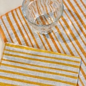 May include: Two yellow and white striped linen kitchen towels. The towels are folded and layered on top of each other. A clear glass cup is sitting on top of the towels.