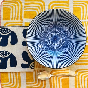 Op de afbeelding: Een blauwe en witte gestreepte keramische kom op een gele en witte gepatroneerde stoffen placemat met een zilveren lepel.