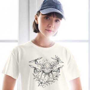 以下が含まれることがあります： クリーム色のTシャツ。黒と白で描かれたドローンの詳細なイラストが、花の要素に囲まれています。デザインにはプロペラ、装飾的な装飾、花が含まれており、芸術的で技術的な融合を生み出しています。Tシャツは、クラシックなクルーネックです。