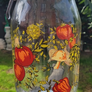 Op de afbeelding: Een glazen voorraadpot met een metalen beugelsluiting en een oranje rubberen ring. De buitenkant is voorzien van een kleurrijke botanische print van rode tomaten, gele bloemen, een beige paddenstoel en groene bladeren met de tekst Made in France.