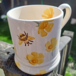 May include: White ceramic mug with a cream-colored interior, adorned with yellow floral and bee designs. The mug has a curved handle and a slightly flared base. The floral pattern includes various sizes of yellow flowers and a single bee. The mug is sitting on a wooden surface.
