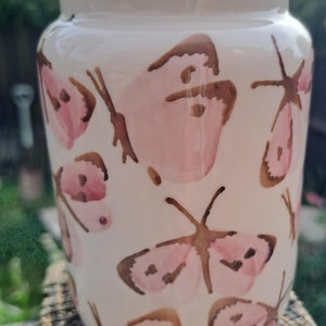 May include: A white ceramic jar with a slightly flared rim, adorned with pink and brown butterfly illustrations. The jar has a cylindrical shape and is suitable for holding small items or as a decorative piece. The butterflies are painted in a watercolor style.