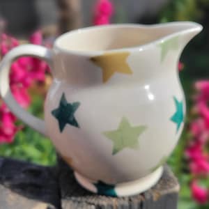 May include: A cream-coloured ceramic jug featuring a rounded body, a curved handle, and a pouring spout. The exterior surface is decorated with a repeating pattern of five-pointed stars in various shades of gold, dark green, and light green.