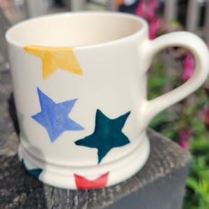 Puede incluir: Una taza de cerámica de color crema con un patrón pintado a mano de estrellas de cinco puntas en varios colores, incluyendo amarillo, azul, verde oscuro y rojo. La taza tiene un asa redondeada y una base ligeramente ensanchada.
