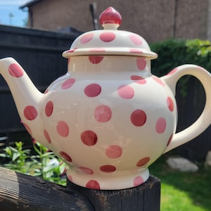 Puede incluir: Una tetera de cerámica de color crema con un patrón de lunares rosas redondos. La tetera tiene un asa curva, un pico vertedor y una tapa extraíble con un pequeño pomo rosa en la parte superior. El diseño es uniforme en todo el cuerpo.