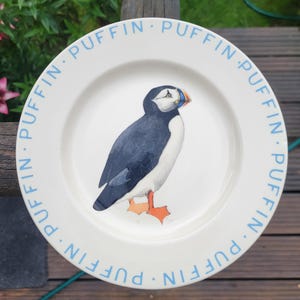 May include: A round white ceramic plate featuring a central illustration of a puffin with black, white, and orange plumage. The rim of the plate is decorated with the word PUFFIN repeated in a light blue sans-serif font around the entire edge.