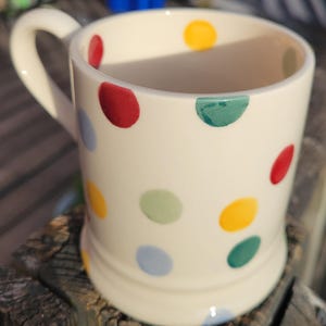 May include: A cream-colored ceramic mug featuring a handle on the left side and a pattern of scattered polka dots in various colors including red, yellow, light blue, and green. The mug has a smooth, cylindrical body with a slightly flared base.