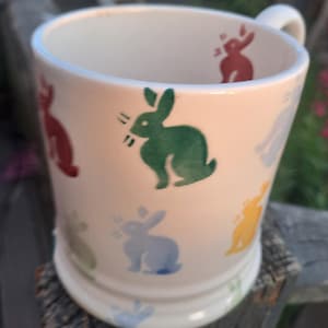 May include: A cream-coloured ceramic mug featuring a repeating pattern of small, stylised rabbits in various colours including green, blue, yellow, and red. The mug has a cylindrical body with a slightly flared base and a white handle on the side.