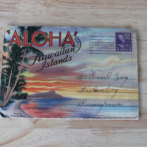 May include: Vintage postcard with the word "ALOHA" and "Hawaiian Islands" in script. The postcard features a sunset scene with palm trees, a beach, and a mountain silhouette. A postage stamp and handwritten address are visible.