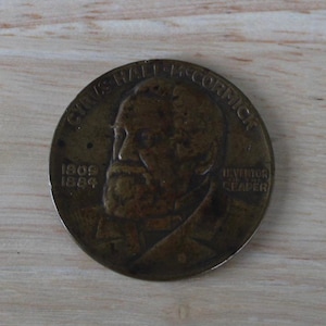 May include: A bronze-colored commemorative coin featuring a portrait of Cyrus Hall McCormick. The coin has the text "CYRUS HALL MCCORMICK" around the top edge, with the years "1809" and "1884" and the words "INVENTOR OF REAPER" visible.