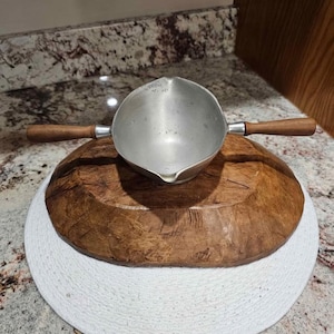 May include: A small, silver-colored saucepan with a wooden handle is placed on top of a large, round, wooden bowl. The bowl sits on a white, woven placemat. The background features a speckled countertop.