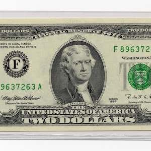 May include: A United States two-dollar bill, featuring a portrait of Thomas Jefferson. The bill is green and black, with the text "TWO DOLLARS" and the serial number "F 89637263 A" visible. The bill is encased in a clear protective sleeve.