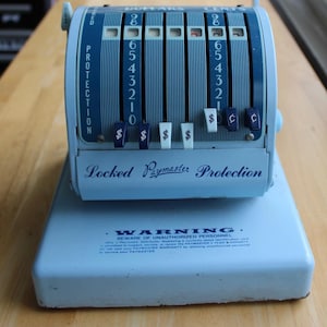 May include: A vintage blue Paymaster cash register with dollar and cent indicators. The register has a series of numbered dials and levers, with the text "Locked Paymaster Protection" and "Warning" visible.
