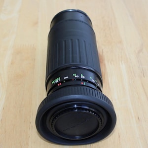 May include: A black camera lens with a ribbed barrel design. The lens has a focus ring with distance markings in feet and meters. The lens cap is also black and has text on it. The lens is on a wooden surface.