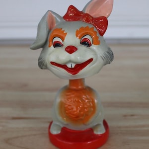 May include: A vintage bobblehead figurine of a smiling rabbit with a red bow, nose, and base. The rabbit has gray fur, orange accents around the eyes, and a cheerful expression. The figurine is a collectible item.