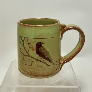 May include: A handmade ceramic mug with a light green exterior and a brown handle. The mug features a framed illustration of a crow perched on a branch. The artist's signature is visible on the side of the mug.