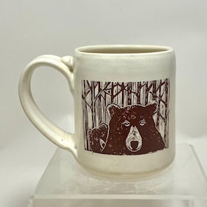 May include: Cream-colored ceramic mug featuring a brown illustration of two bears in a forest. The mug has a curved handle and a cylindrical shape. The bear design is a linocut style.