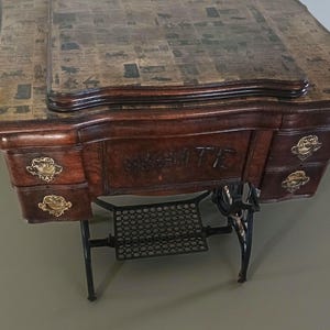 May include: Antique sewing machine table with a dark wood finish. The tabletop is covered in a newspaper print design. The table has drawers with ornate gold-colored hardware and the word "WHITE" is carved into the front.