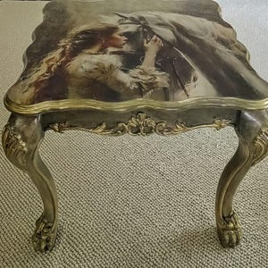 May include: A square side table with a scalloped edge and cabriole legs featuring carved claw feet. The table surface displays a printed image of a person with long hair touching the face of a white horse. The frame is finished in metallic gold.