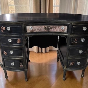 May include: A black kidney-shaped desk featuring a central drawer with a marbled pattern and eight side drawers. Each drawer is accented with clear crystal-style knobs and small floral decals. The desk stands on four slender, tapered legs.