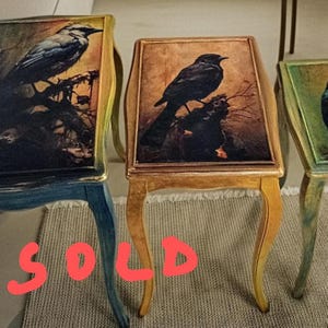 May include: A set of three nesting tables with curved legs, each featuring a rectangular top decorated with a detailed bird illustration. The largest table is blue, the middle is gold, and the smallest is green, with the word SOLD in red text.