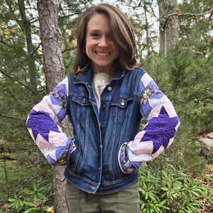 May include: A dark wash denim jacket with quilted sleeves. The sleeves feature a patchwork design with purple star shapes, pink, and floral patterns. The jacket has a classic collar and button-down front.