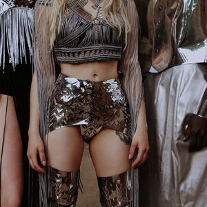 May include: A fashion ensemble featuring a silver metallic crop top, skirt, and thigh-high boots. The outfit is paired with a fringed shawl. The overall aesthetic is futuristic and avant-garde.