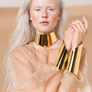 May include: A woman with long, silver hair wears a gold collar necklace and matching cuff bracelets. The jewelry is highly reflective, and the woman is wearing a sheer, beige top. The background is a neutral beige.