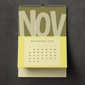 May include: A wall calendar for November 2026, featuring a large "NOV" in a light color against a green background. The calendar includes a yellow section with the month's dates, and a cream-colored bottom section. The calendar is hanging on a wall.