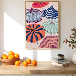 May include: A framed art print featuring colorful, abstract umbrella-like shapes in shades of orange, blue, pink, and green. The artwork has a light background and is displayed on a wall. A wooden bowl of oranges sits on a wooden surface in the foreground.
