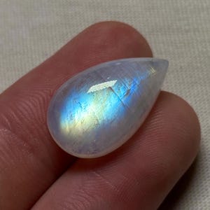 May include: A teardrop-shaped moonstone gemstone with an iridescent sheen. The stone exhibits a play of colour, with blues, yellows, and whites visible. The polished surface reflects light, creating a shimmering effect. The gemstone is held between fingers.
