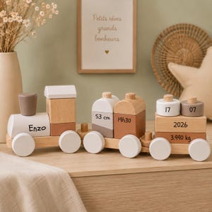 Könnte beinhalten: Ein hölzerner Spielzeugeisenbahnzug mit drei verbundenen Wagen in Weiß-, Beige- und Brauntönen. Die Lokomotive ist mit Enzo beschriftet. Der mittlere Wagen zeigt 53 cm und 19h30. Der hintere Wagen trägt die Zahlen 17, 07, 2026 und 3.990 kg.