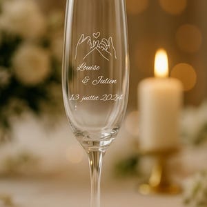 Puede incluir: Una copa de champán transparente con un delicado diseño de manos entrelazadas y un corazón. Los nombres "Louise & Julien" y la fecha "13 juillet 2024" están grabados en el cristal. Se ve una vela encendida y un fondo borroso.