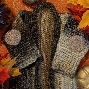 Könnte beinhalten: Ein handgefertigter Baby-Pullover in Grau-, Braun- und Schwarztönen, mit Ellbogenpatches und einem strukturierten Design. Der Pullover wird mit herbstlichen Dekorationen wie Kürbissen und Blättern präsentiert. Der Text auf dem Bild lautet "Lil' toasted mallow Elbow Patch Baby Sweater."