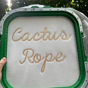 May include: A white fabric with the words "Cactus Rope" embroidered in a rope-like font, held within a green embroidery hoop. The embroidery is in a light tan color.