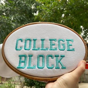 Puede incluir: Un bastidor de bordado ovalado con las palabras "COLLEGE BLOCK" bordadas en hilo turquesa sobre tela blanca. El bastidor de madera se sostiene en una mano, con un fondo verde borroso.